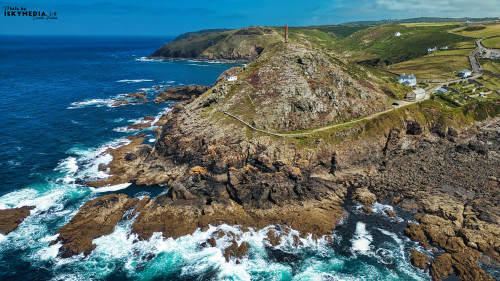 Aerial view of a rugged coastline with rocky cliffs and crashing waves. A lighthouse stands on top of a hill, surrounded by green fields and a winding road. The ocean is a deep blue, contrasting with the rocky shore.