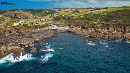 A scenic coastal view featuring rocky shores, clear blue waters, and green hills in the background. The landscape includes a small village with white houses and a winding path leading to the shore, under a partly cloudy sky.