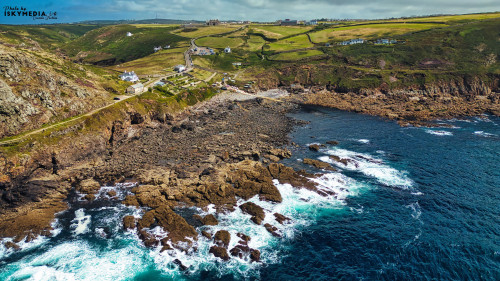 Aerial view of a rugged coastline with rocky shores and crashing waves. Lush green hills rise above the sea, dotted with white houses. The sky is partly cloudy, creating a picturesque landscape.