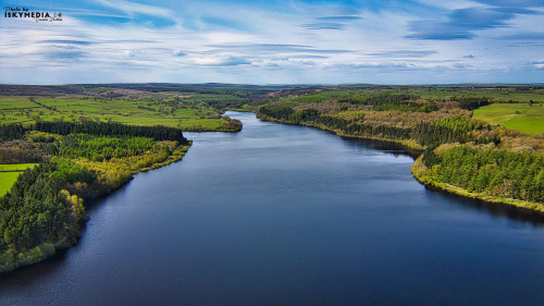 Aerial view of a serene lake surrounded by lush forests and open fields under a partly cloudy sky.