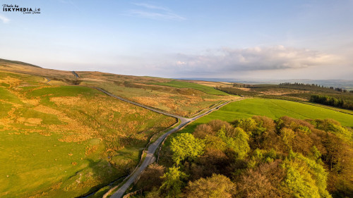 Aerial view of a winding road through lush green countryside with hills under a clear sky.