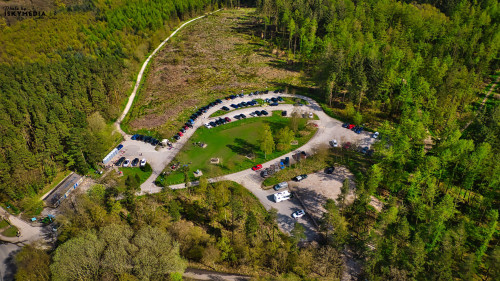 Aerial view of a curved parking area surrounded by lush green forests, with cars parked and a dirt road leading into the woods.