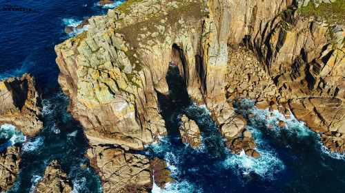 Aerial view of rugged coastal cliffs with a natural rock arch, surrounded by deep blue ocean waters. Waves crash against the rocky shoreline, creating a dynamic seascape. The cliffs are textured with moss and lichen, showcasing natural beauty.