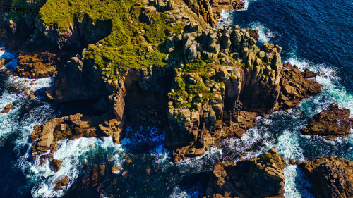 Aerial view of rugged coastal cliffs surrounded by deep blue ocean waves. The rocky formations are covered with green vegetation, creating a striking contrast against the water. The scene captures the beauty of nature and the power of the sea.