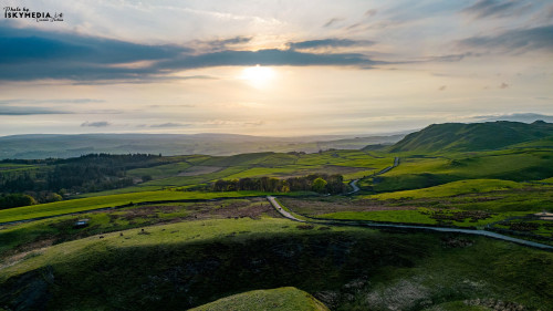 Scenic sunset over rolling hills with a winding road, lush greenery, and a tranquil sky.