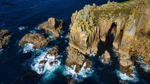 Aerial view of rugged coastal cliffs and rocky formations surrounded by deep blue ocean waves. The cliffs are adorned with patches of green moss and lichen, creating a striking contrast against the water. The scene captures the beauty of nature's coastline.
