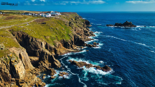 A stunning aerial view of a rugged coastline with cliffs, lush green hills, and the deep blue ocean. The scene features a small village or settlement perched on the cliffs, with waves crashing against the rocks below. The sky is clear with a few clouds, enhancing the beauty of the landscape.
