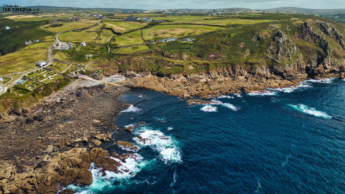 Aerial view of a rugged coastline with rocky shores and crashing waves. Lush green fields and a winding road are visible on the land, leading to a small settlement. The ocean is deep blue, contrasting with the rocky landscape.