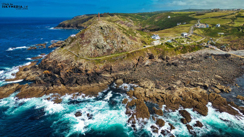 Aerial view of a rugged coastline featuring rocky cliffs, crashing waves, and a lighthouse on a hill. The landscape is lush and green, with a winding road and scattered white houses in the background. The ocean is a vibrant blue, contrasting with the rocky shore.