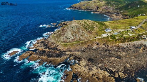 Aerial view of a coastal landscape featuring rocky shores, a lighthouse on a hill, and lush green hills in the background. The ocean waves crash against the rocks, creating a scenic and tranquil atmosphere.