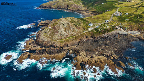 Aerial view of a rugged coastline featuring rocky cliffs, crashing waves, and a lighthouse on a hill. The landscape includes green fields and a small village, with the ocean in vibrant shades of blue and turquoise.