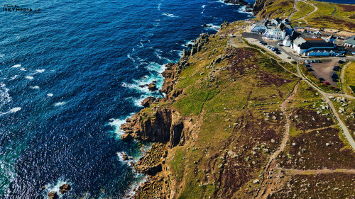 Aerial view of a rugged coastline with cliffs and a blue ocean. A small village is visible near the shore, surrounded by green and brown hills. Waves crash against the rocks, creating a scenic landscape.