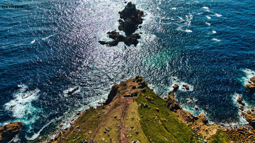 Aerial view of a rocky coastline with a small island in the ocean. The water is shimmering under sunlight, creating a beautiful contrast with the green land and rocky formations. Waves gently crash against the shore, enhancing the natural beauty of the scene.