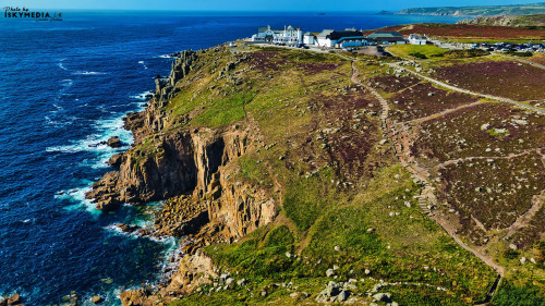Aerial view of a rugged coastline with cliffs and a hotel on top. The landscape features green hills, rocky outcrops, and the deep blue ocean. The sky is clear, and the scene captures the natural beauty of the coastal area.
