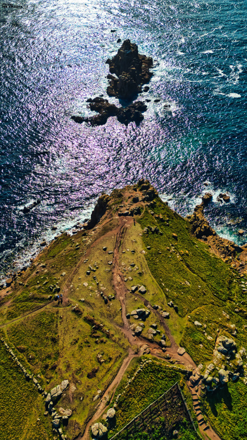 Aerial view of a coastal landscape featuring rocky formations and lush greenery. The shimmering ocean reflects sunlight, creating a scenic contrast with the rugged terrain. Paths wind through the landscape, leading to the rocky outcrop in the distance.
