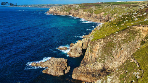 A stunning coastal view featuring rugged cliffs and clear blue waters. The landscape showcases a mix of rocky formations and lush greenery, with waves gently crashing against the shore. The horizon reveals a distant coastline under a clear sky.