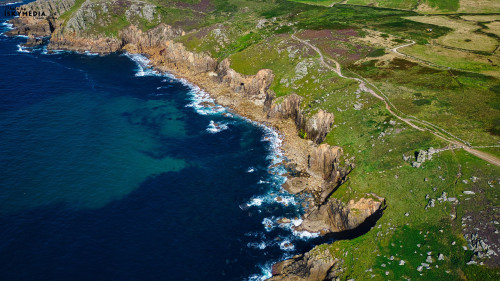 Aerial view of a rugged coastline with cliffs, green hills, and blue ocean waves crashing against the rocks. The landscape features a mix of rocky terrain and lush vegetation, showcasing the natural beauty of the area.