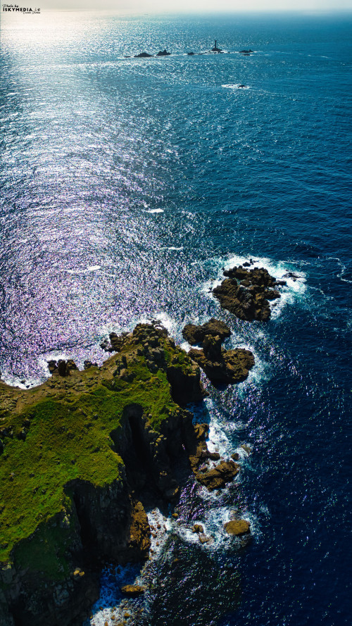Aerial view of a rocky coastline with lush green vegetation, surrounded by deep blue ocean waters. The sunlight reflects off the water, creating a shimmering effect. In the distance, a lighthouse can be seen on a small island.