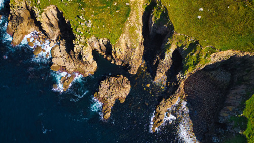Aerial view of rugged coastal cliffs meeting the ocean. The landscape features rocky formations, lush green vegetation, and waves crashing against the shore. The contrast between the dark blue water and the bright green land creates a stunning natural scene.
