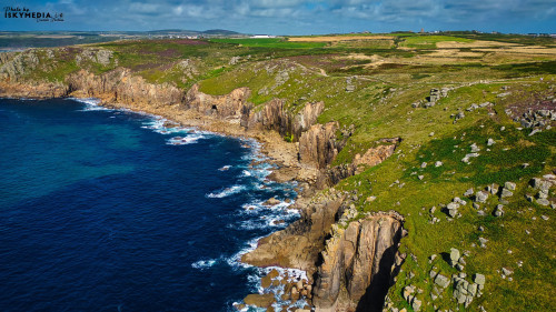 Aerial view of rugged coastal cliffs meeting the deep blue sea, with lush green fields and rolling hills in the background. The coastline features rocky formations and waves crashing against the shore under a partly cloudy sky.