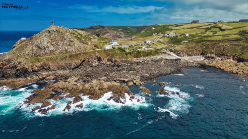A scenic coastal landscape featuring rocky shores, rolling hills, and a lighthouse on a hilltop. The ocean waves crash against the rocks, with a clear blue sky and scattered clouds in the background. A small village is visible near the coastline.