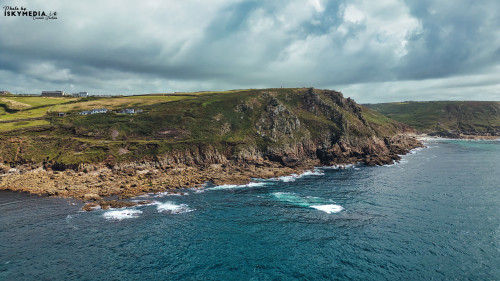 A scenic coastal view featuring rugged cliffs and rocky shorelines. The ocean waves crash against the rocks, with green hills in the background under a cloudy sky. This picturesque landscape captures the beauty of nature.