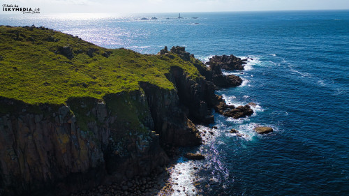 Aerial view of rugged coastal cliffs meeting the ocean, with waves crashing against the rocks. The scene features lush green vegetation on the cliffs and distant rock formations in the water, under a bright blue sky.