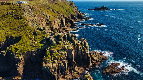 Aerial view of rugged coastal cliffs with lush greenery, overlooking the deep blue ocean. The landscape features rocky formations and a distant island, creating a dramatic seascape under clear skies.