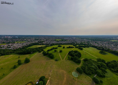 Aerial view of a lush green park with walking paths, surrounded by a suburban landscape under a hazy sky in London.