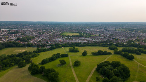 Aerial view of a suburban landscape with green parks and residential areas under a hazy sky in London.