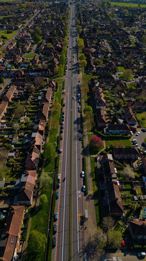 Aerial shot of a long straight road cutting through a residential area lined with houses, cars, and trees in Morden, London, UK.