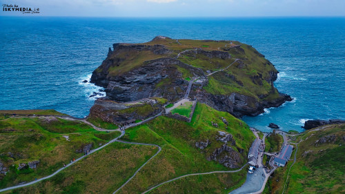 Aerial view of a rugged coastal landscape featuring a rocky island surrounded by turquoise waters. The island has winding paths and green hills, with a small building at the base. The sky is overcast, adding a dramatic effect to the scenery.