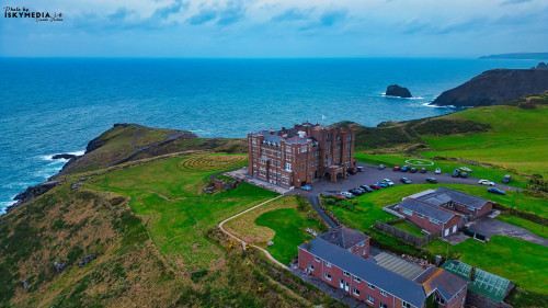 Aerial view of a large historic building situated on a cliff overlooking the ocean. The structure features multiple stories and is surrounded by green fields and a parking area. The coastline is rugged with rocky formations and a cloudy sky above.