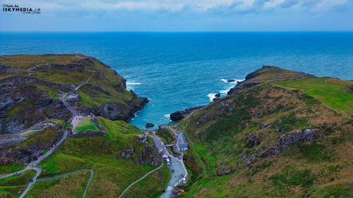 Aerial view of a coastal landscape featuring rugged cliffs, a winding road, and the ocean in the background. The scene captures the natural beauty of the coastline with green hills and rocky formations.