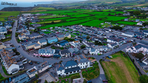 Aerial view of a coastal village with green fields, parked cars, and a winding road. The village features a mix of traditional and modern buildings, with a backdrop of hills and cliffs. The scene captures the charm of rural life near the coast.