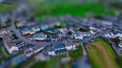 Aerial view of a small town with winding streets, featuring a mix of residential and commercial buildings. The scene captures the charm of a quaint village, surrounded by green fields.