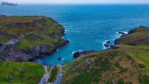 Aerial view of a coastal landscape featuring rugged cliffs, lush green hills, and a serene blue ocean. The coastline is dotted with rocky outcrops and winding paths, showcasing the natural beauty of the area.