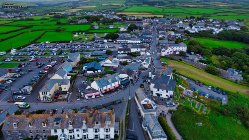 Aerial view of a small town with a mix of residential and commercial buildings. The town features a winding road, parked cars, and green fields in the background. The architecture includes traditional houses and shops, with a parking area visible.