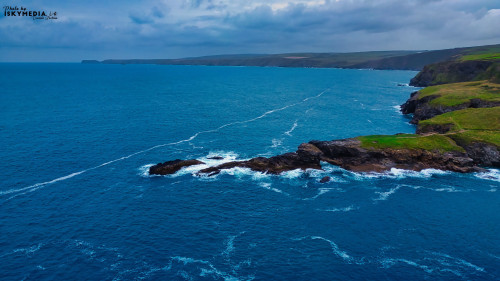 Aerial view of a rugged coastline with rocky outcrops and waves crashing against them. The ocean is a deep blue, contrasting with the green hills in the background under a cloudy sky.