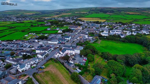 Aerial view of a small village surrounded by lush green fields. The village features a mix of residential and commercial buildings, with a parking area visible. The landscape includes rolling hills and farmland in the background, creating a picturesque rural scene.
