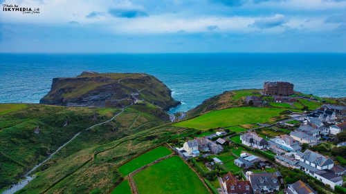 Aerial view of a coastal landscape featuring a rocky outcrop, lush green hills, and a small village by the sea. The ocean is visible in the background, with a cloudy sky overhead.