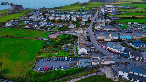 Aerial view of a coastal village with a mix of residential and commercial buildings. The landscape features green fields, parked cars, and a road leading to the sea. The ocean is visible in the background, with a few boats on the water.