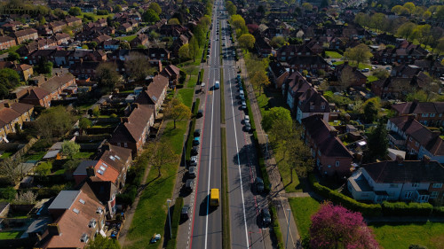Aerial shot of a residential street lined with houses, cars, and trees. A multi-lane road runs through the center.