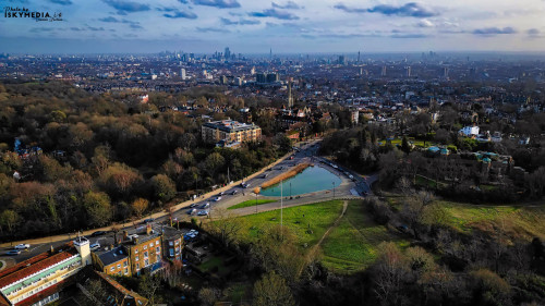 Aerial view of a city park with a pond, roads, and distant skyline at dusk.