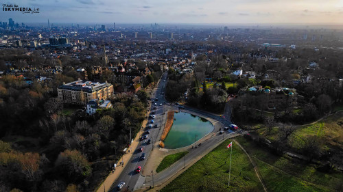 Aerial view of a city park at dusk with roads and a pond, urban skyline in the distance.