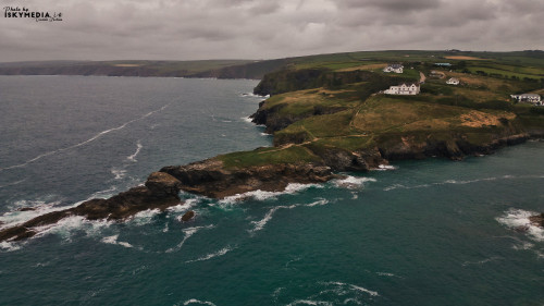 A scenic coastal view featuring rugged cliffs, rolling green hills, and white houses along the shoreline. The ocean waves crash against the rocky outcrops under a cloudy sky, creating a dramatic atmosphere.
