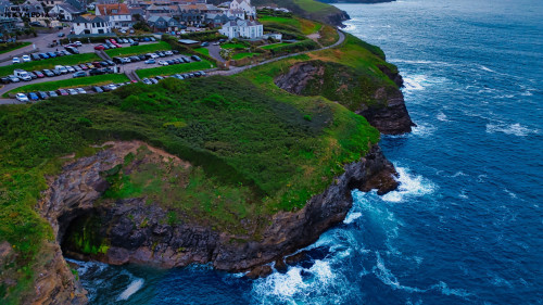 Aerial view of a coastal landscape featuring cliffs, waves crashing against rocks, and a small village with houses and parked cars. The lush green hills contrast with the blue ocean, creating a picturesque scene.