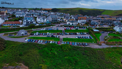 Aerial view of a coastal village with a large parking area filled with cars. The surrounding landscape features green hills and a mix of residential buildings, showcasing a picturesque seaside setting.