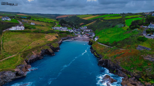 Aerial view of a coastal village surrounded by lush green hills and rocky shores. The turquoise water contrasts with the dark rocks and sandy beach. White houses dot the landscape, creating a picturesque scene of nature and tranquility.