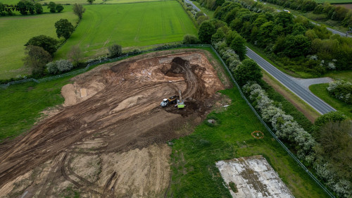 Excavator and truck working on a large dirt area surrounded by green fields and trees at Bellway Homes in Boroughbridge, UK.